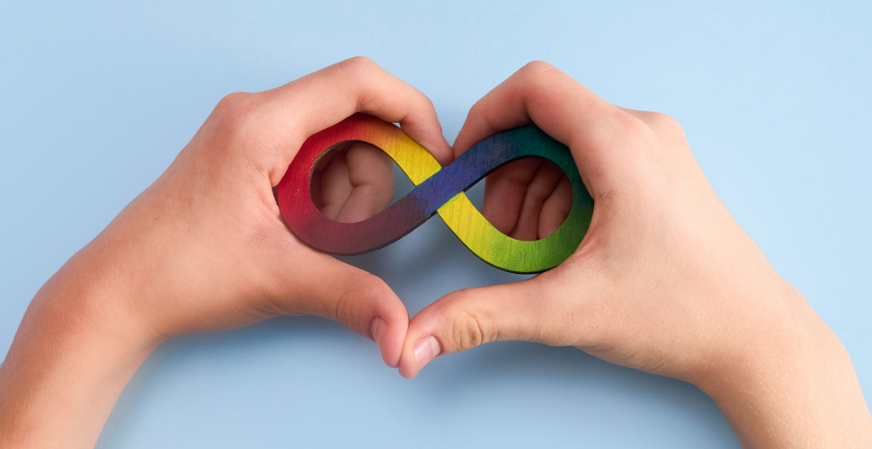 Hands forming a heart shape around a rainbow-coloured infinity symbol, representing neurodiversity and inclusion.