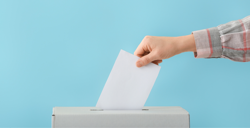 A person placing a ballot into a box against a blue background, symbolising civic engagement, active participation, and the courage to make every voice count.
