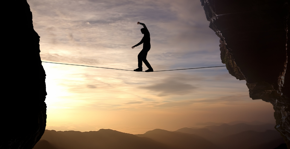 A person walks a tightrope between two cliffs at sunrise, symbolising the quiet struggle and balance required to lead through challenge and change.