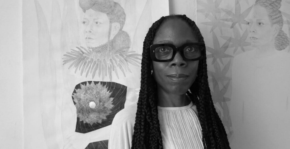 Black and white portrait of artist Charmaine Watkiss standing in front of her artwork, representing creativity, identity and the power of cultural storytelling.