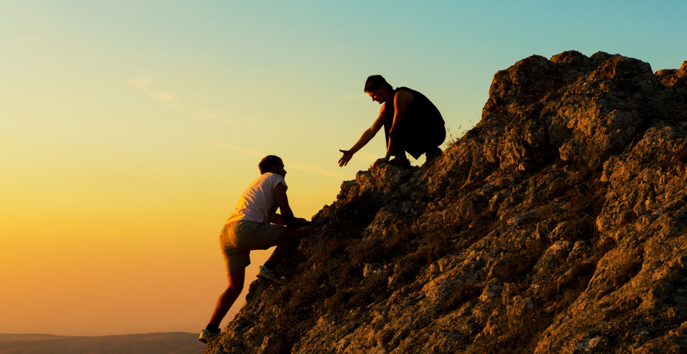 Two people climbing a rocky hill at sunset, with one reaching out to help the other, symbolising teamwork and support.