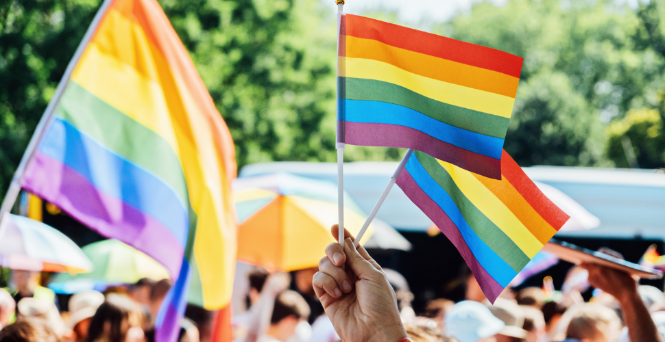 A person holds rainbow Pride flags at a vibrant outdoor celebration, symbolising unity, visibility and the intersection of LGBTQ+ identities.