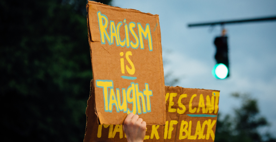A protestor holds a cardboard sign reading “Racism is taught,” symbolising the call to confront and unlearn racial prejudice.
