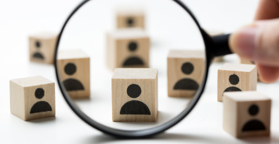 A magnifying glass focusing on a wooden block with a person icon, symbolising inclusive and diverse recruitment for trustee roles.